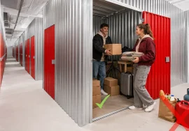 Young couple placing boxes, a suitcase and a bicycle in a City Storage unit, with examples of prohibited food and hazardous materials.