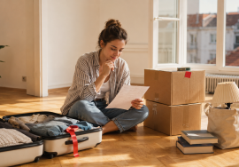 Young woman preparing to move abroad with a suitcase, moving boxes and belongings selected for donation